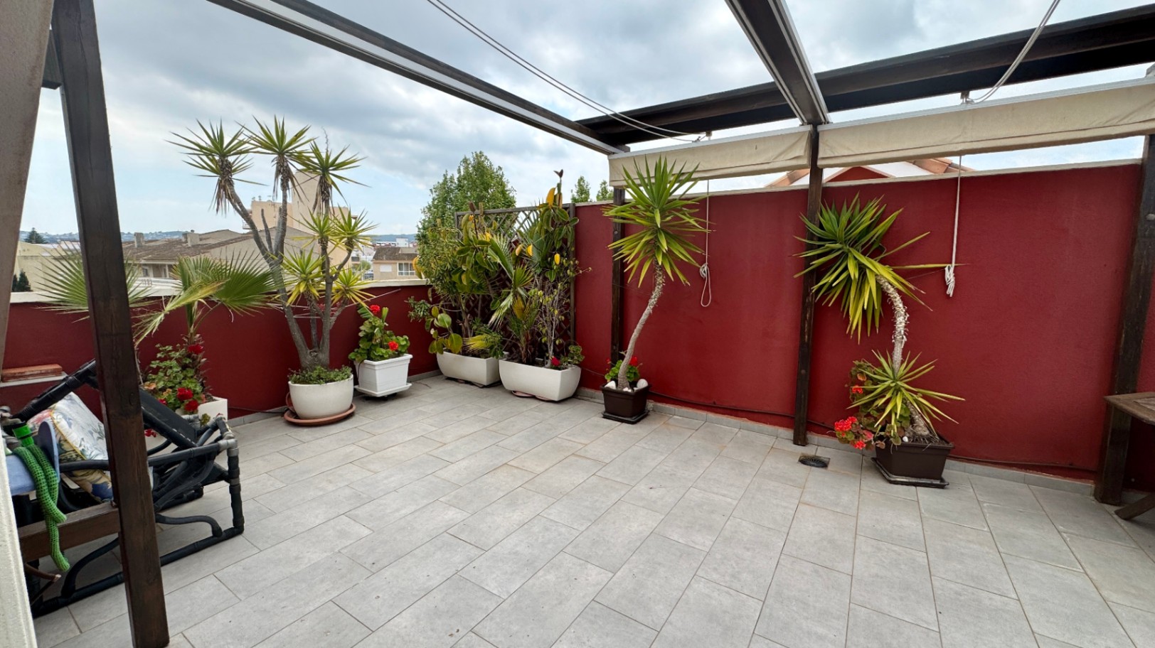 Spacious rooftop terrace featuring grey tiled flooring, a wooden pergola with retractable awnings, and a collection of potted Mediterranean plants against a red accent wall.