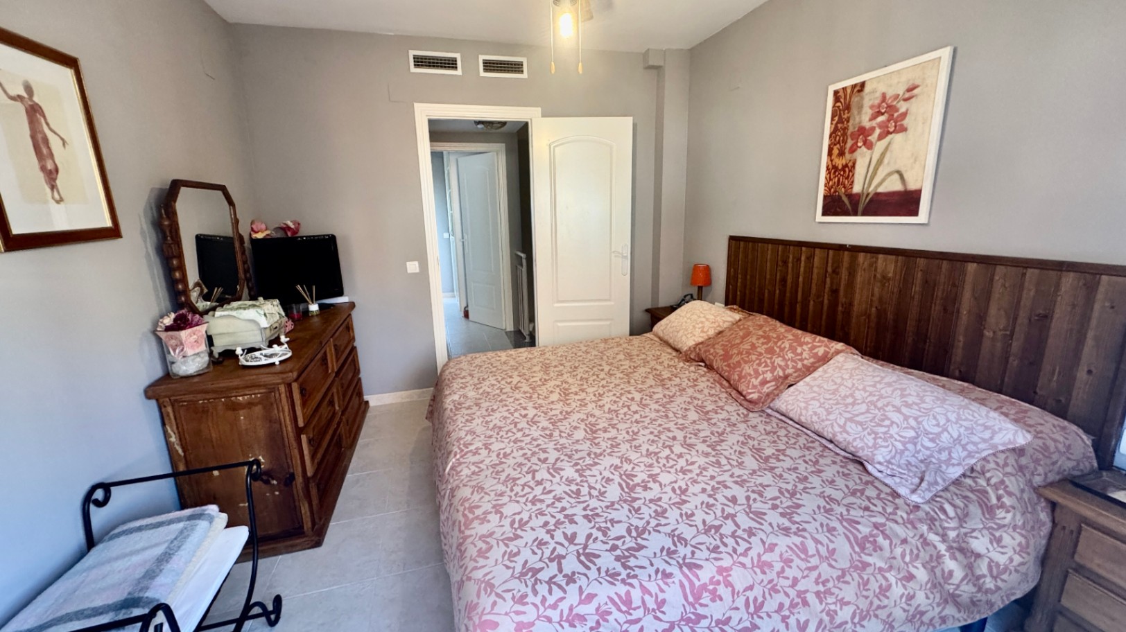 Bedroom featuring a dark wood slat headboard, rustic dresser with mirror, and light grey walls. The space includes tiled flooring and integrated air conditioning vents.