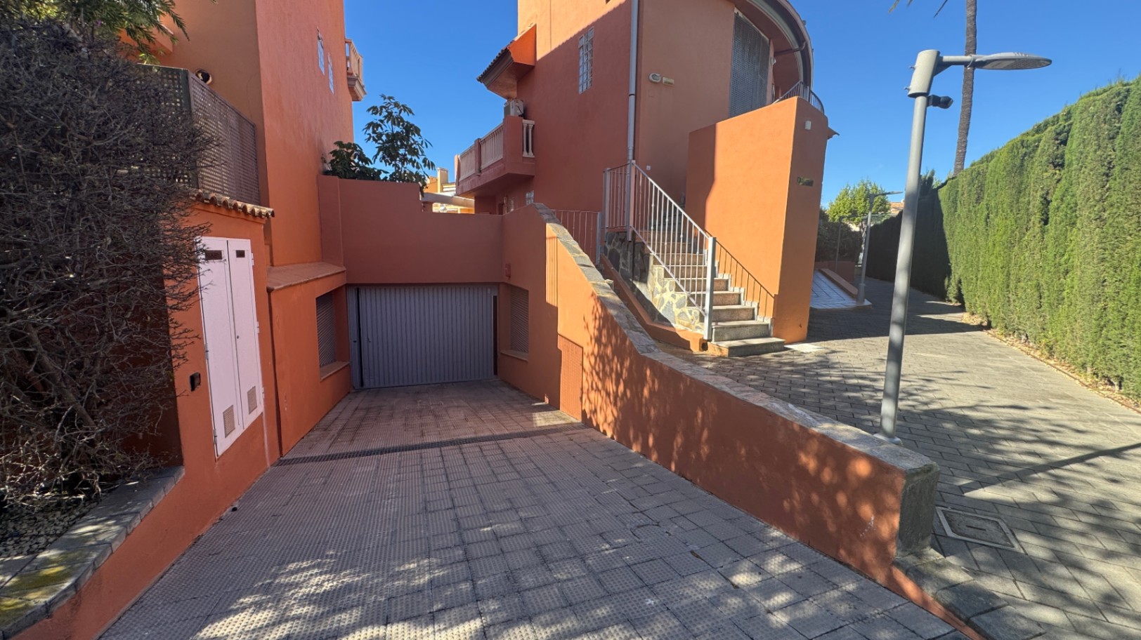 Exterior view featuring a paved driveway leading to a private garage, terracotta-colored facade, and outdoor stairs with metal railings beside a tall hedge.