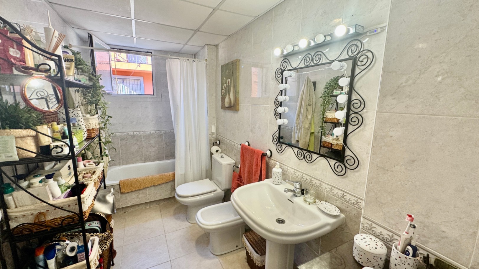 Fully tiled bathroom featuring a pedestal sink, a mirror with decorative ironwork and integrated bulb lighting, a bidet, and a bathtub.