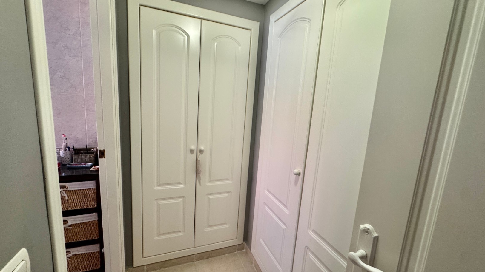 Hallway featuring floor-to-ceiling white built-in wardrobes with arched panel detailing, situated adjacent to a bathroom with wicker storage.