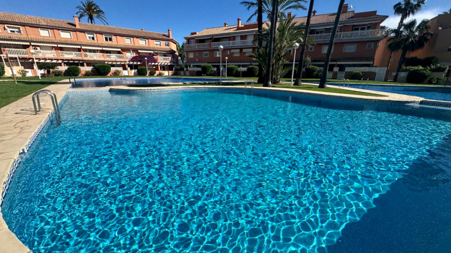 Large communal swimming pool with blue mosaic tiling and a stone border, surrounded by landscaped gardens and Mediterranean-style buildings.