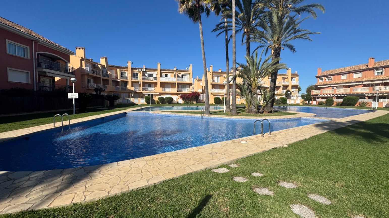 Large communal swimming pool with blue tiling, surrounded by a stone-paved sun deck, manicured lawns, and palm trees within a residential complex.