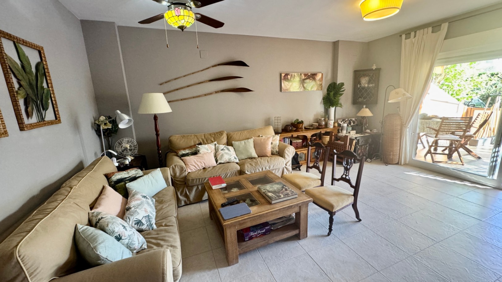 Living area featuring beige sofas, a rustic wooden coffee table, and large glass doors opening onto a furnished sunlit terrace.