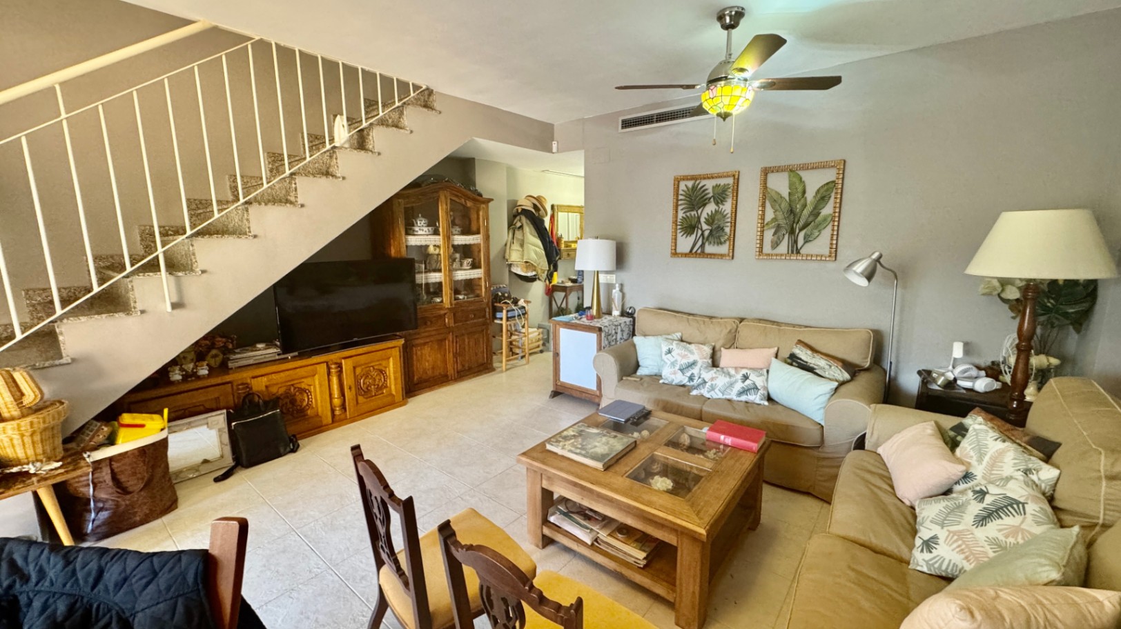 Living area featuring beige sofas, a wooden coffee table, and a staircase with white railings. Includes a wooden TV unit and display cabinet integrated under the stairs.