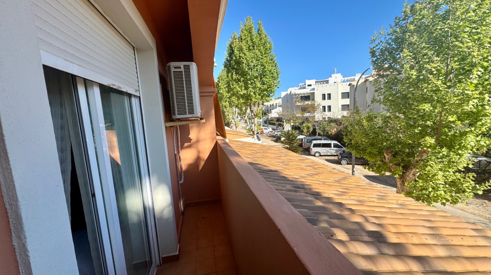 Narrow balcony with terracotta tiling, featuring an external AC unit and views of a tree-lined street with residential buildings.
