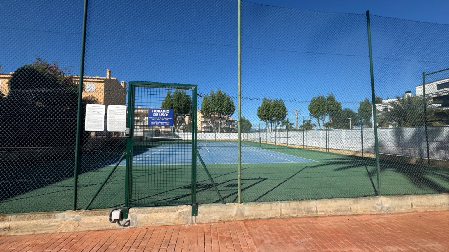 Outdoor tennis court with a blue and green hard surface, enclosed by a high chain-link fence and accessible via a secure green metal gate.