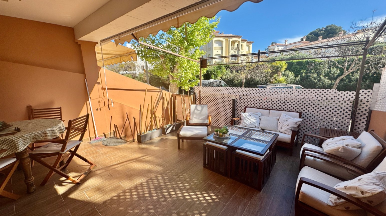 Spacious terrace with wood-effect tiling, featuring a lounge area with white cushions, a wooden dining set, and retractable awnings.