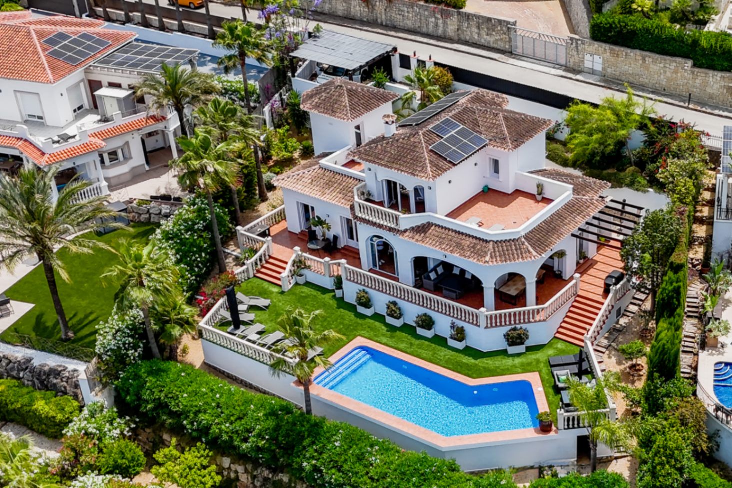 Aerial perspective of a Mediterranean villa with a geometric pool, rooftop solar panels, and multi-level terraces featuring terracotta tiles and white balustrades.