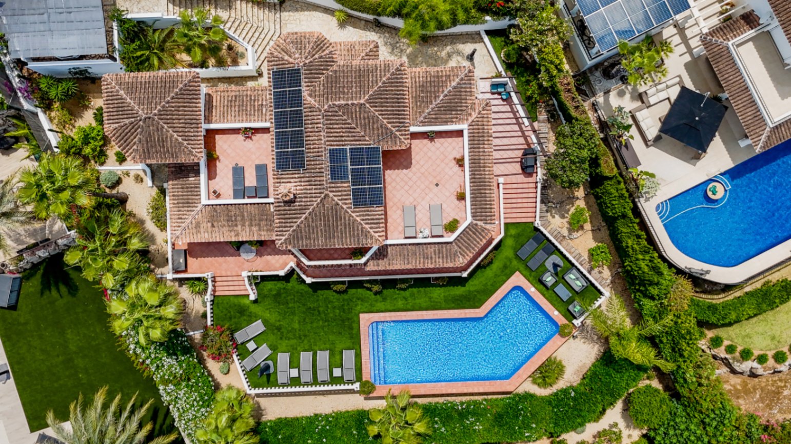 Aerial view of a Mediterranean villa featuring a terracotta tile roof with solar panels, multiple sun terraces, and an angular swimming pool surrounded by manicured lawns.