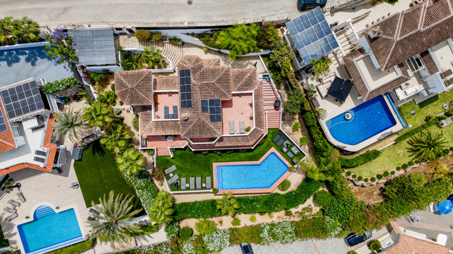 Aerial view of a villa featuring terracotta roofing with solar panels, a geometric swimming pool, and landscaped gardens with palm trees and sun terraces.