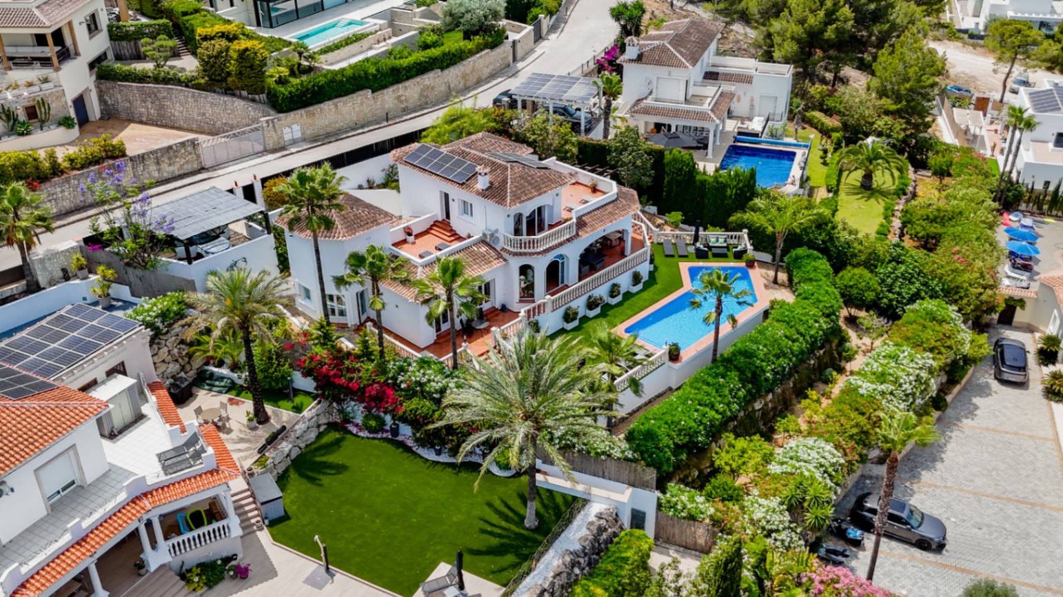 Aerial view of a white Mediterranean villa featuring terracotta roofs, solar panels, a private geometric pool, and landscaped gardens with palm trees.