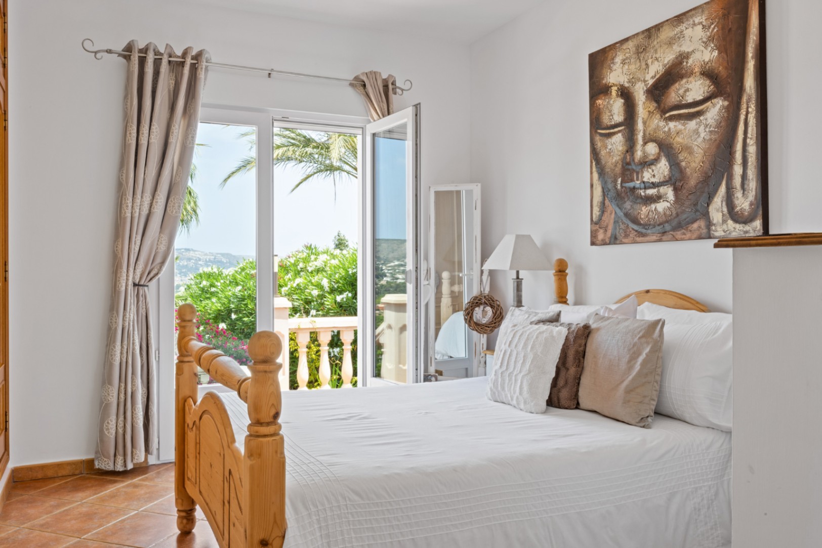 Bedroom featuring a wooden bed frame, terracotta flooring, and a large Buddha artwork. Glass doors open to a balcony with views of Mediterranean greenery and distant hills.