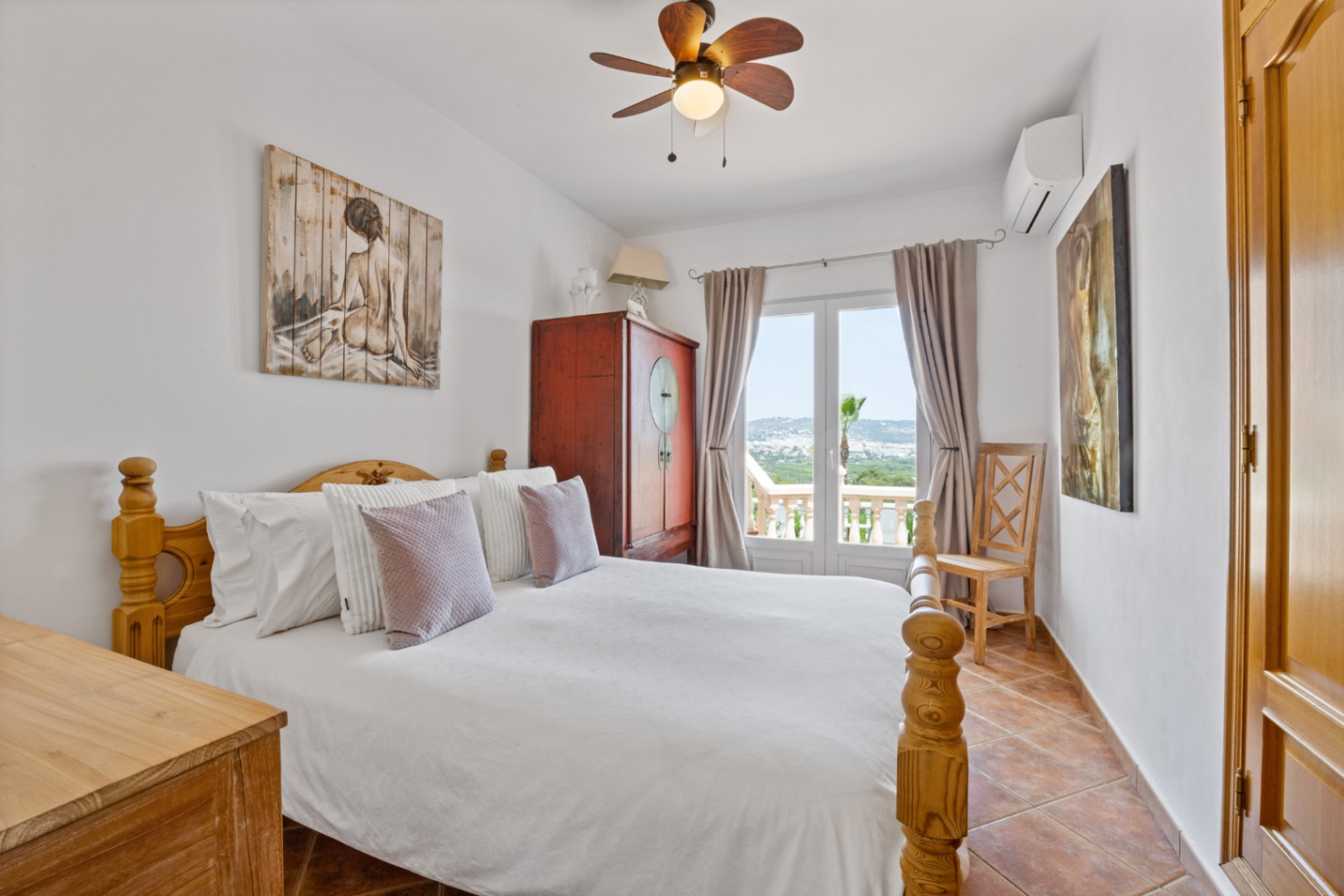 Bedroom featuring a wooden bed frame, terracotta tile flooring, and French doors opening to a balcony with landscape views. Includes a ceiling fan and AC.