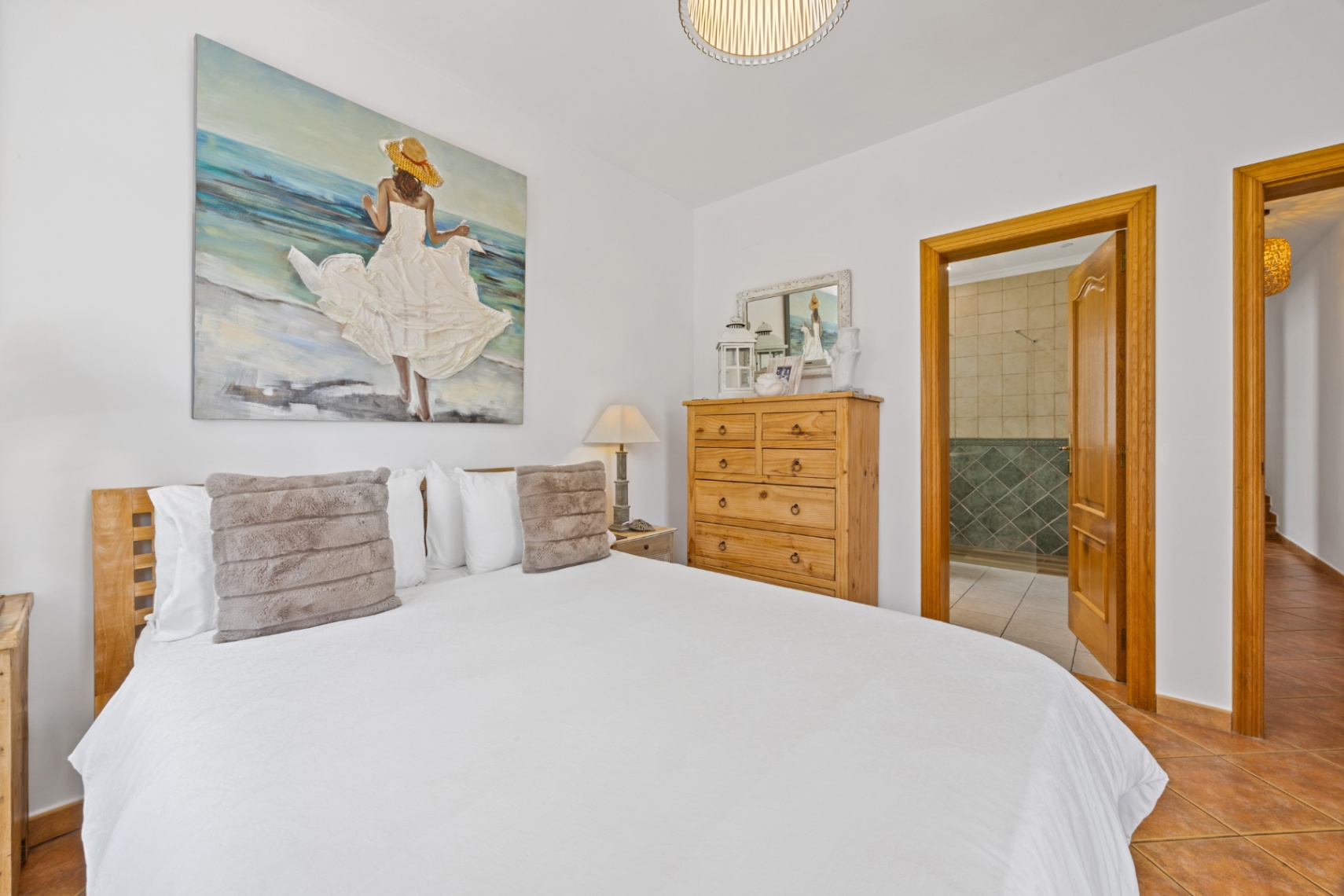 Bedroom featuring a wooden headboard, large coastal-themed artwork, and terracotta tile flooring. An open doorway reveals an ensuite bathroom with green tiled walls.