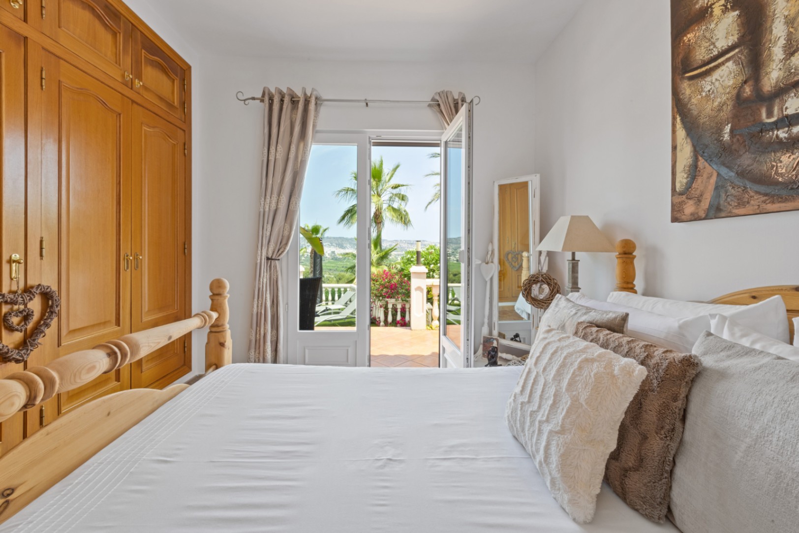 Bright bedroom featuring a wooden bed and built-in wardrobe, with French doors opening onto a terrace with palm trees and landscape views.