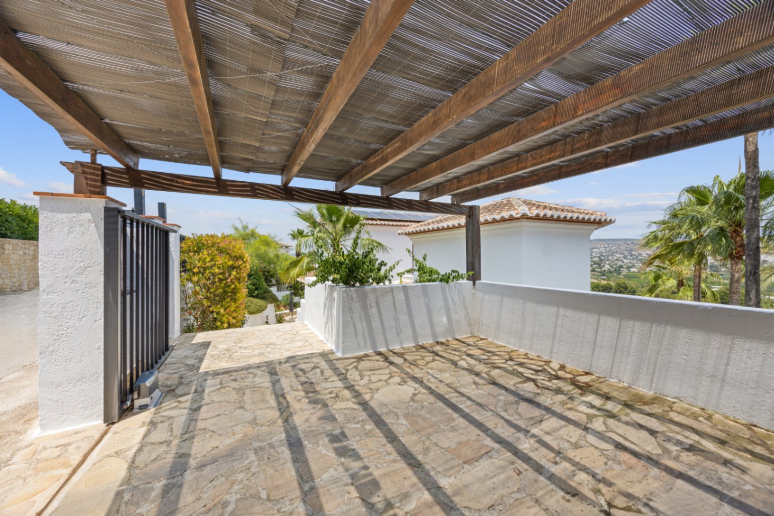 Covered parking area with a wooden pergola and reed roofing, featuring irregular stone flooring and views of the surrounding valley.