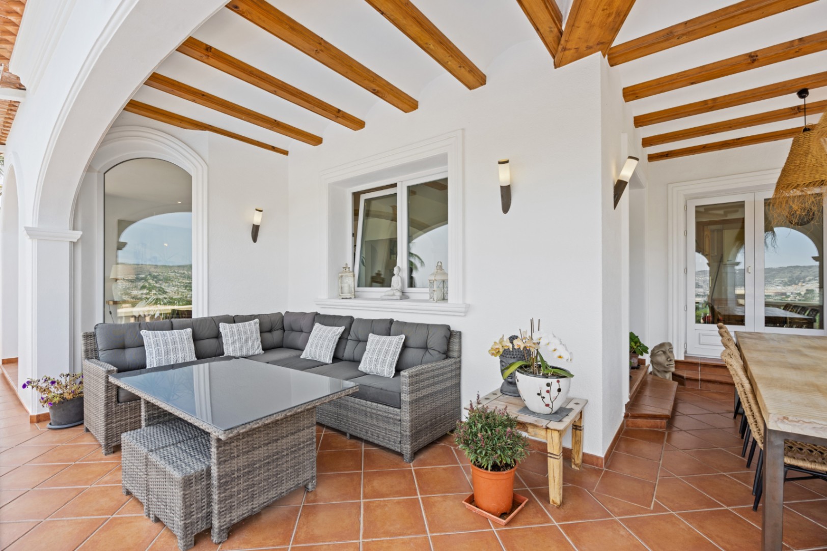 Covered terrace featuring exposed wooden beams, terracotta flooring, and a grey wicker lounge set. The space includes wall-mounted lighting and landscape views.