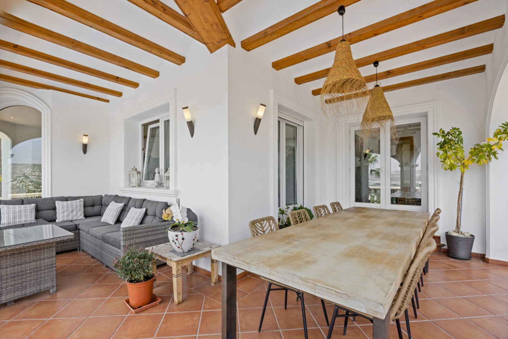 Covered terrace featuring exposed wooden beams, terracotta tiles, a large wooden dining table with woven chairs, and an L-shaped wicker lounge set.
