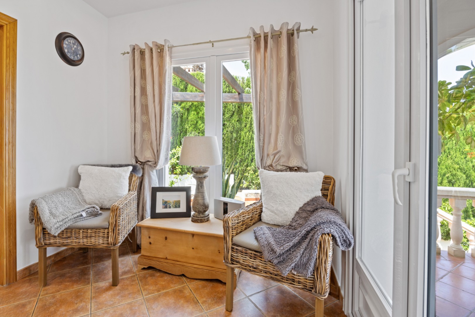 Cozy seating area featuring two wicker armchairs, a rustic wooden chest, and terracotta flooring. Large windows provide natural light and views of the garden.