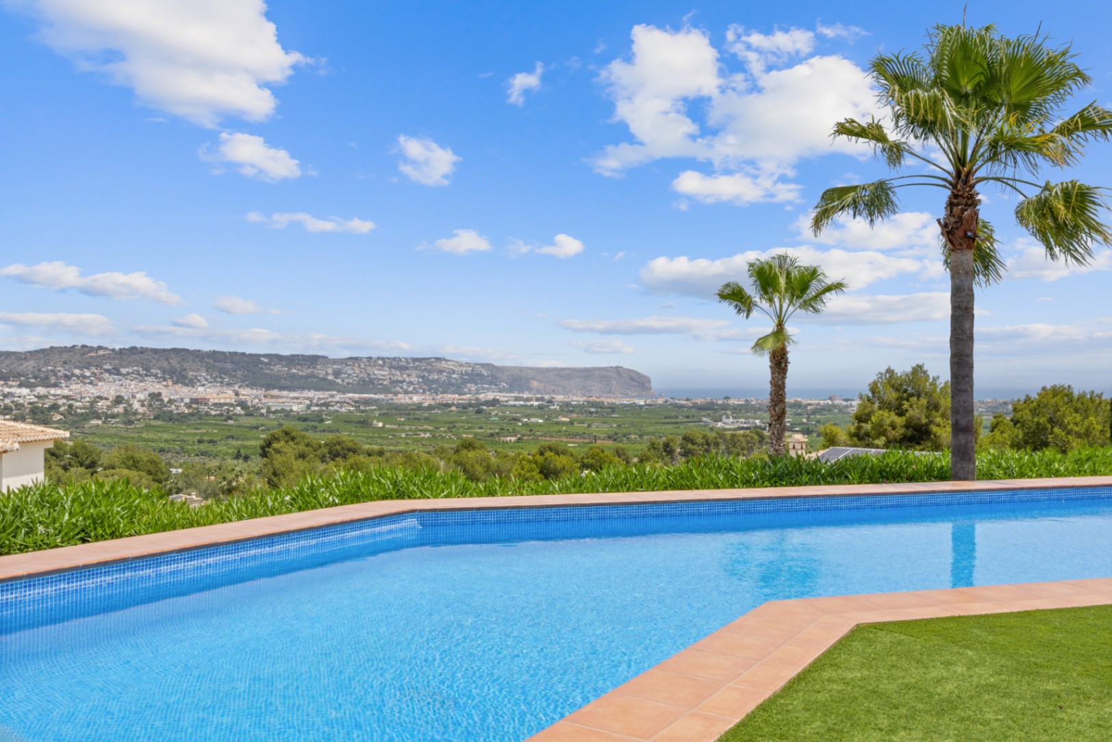 Curved swimming pool with blue mosaic tiles and terracotta coping, offering panoramic views of the valley, town, and distant hills.