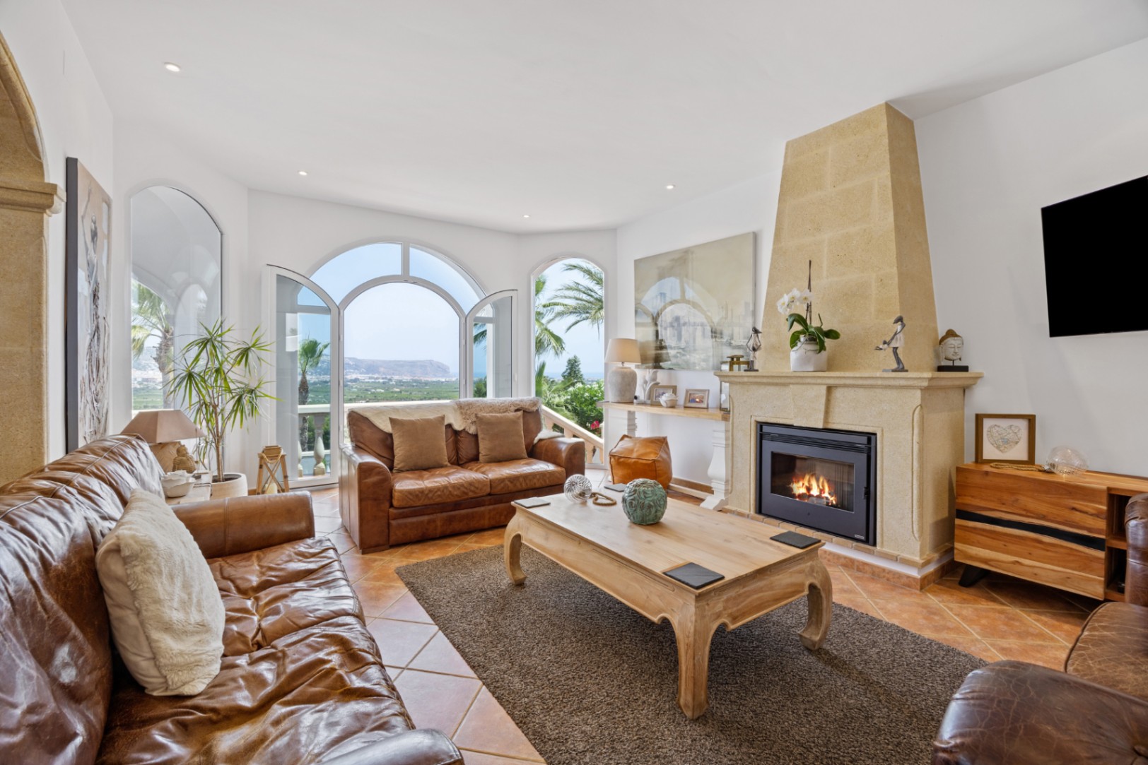 Elegant living room featuring a stone fireplace, brown leather seating, and arched doors opening to a terrace with panoramic sea and mountain views.
