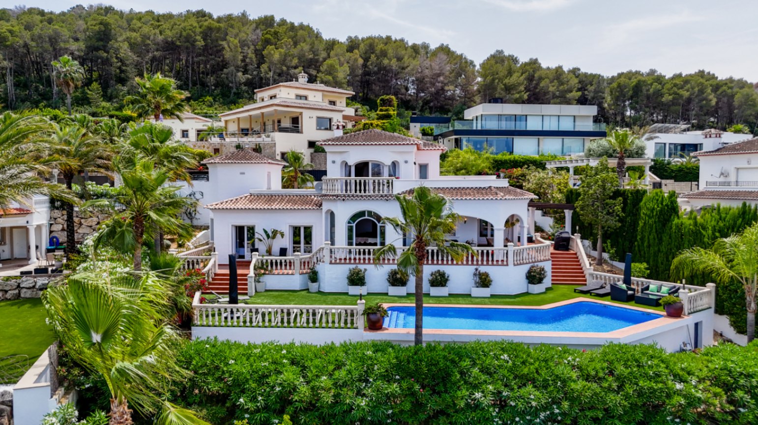 Elevated view of a white Mediterranean villa featuring terracotta roofs, arched windows, and a private pool surrounded by palm trees and pine forest.