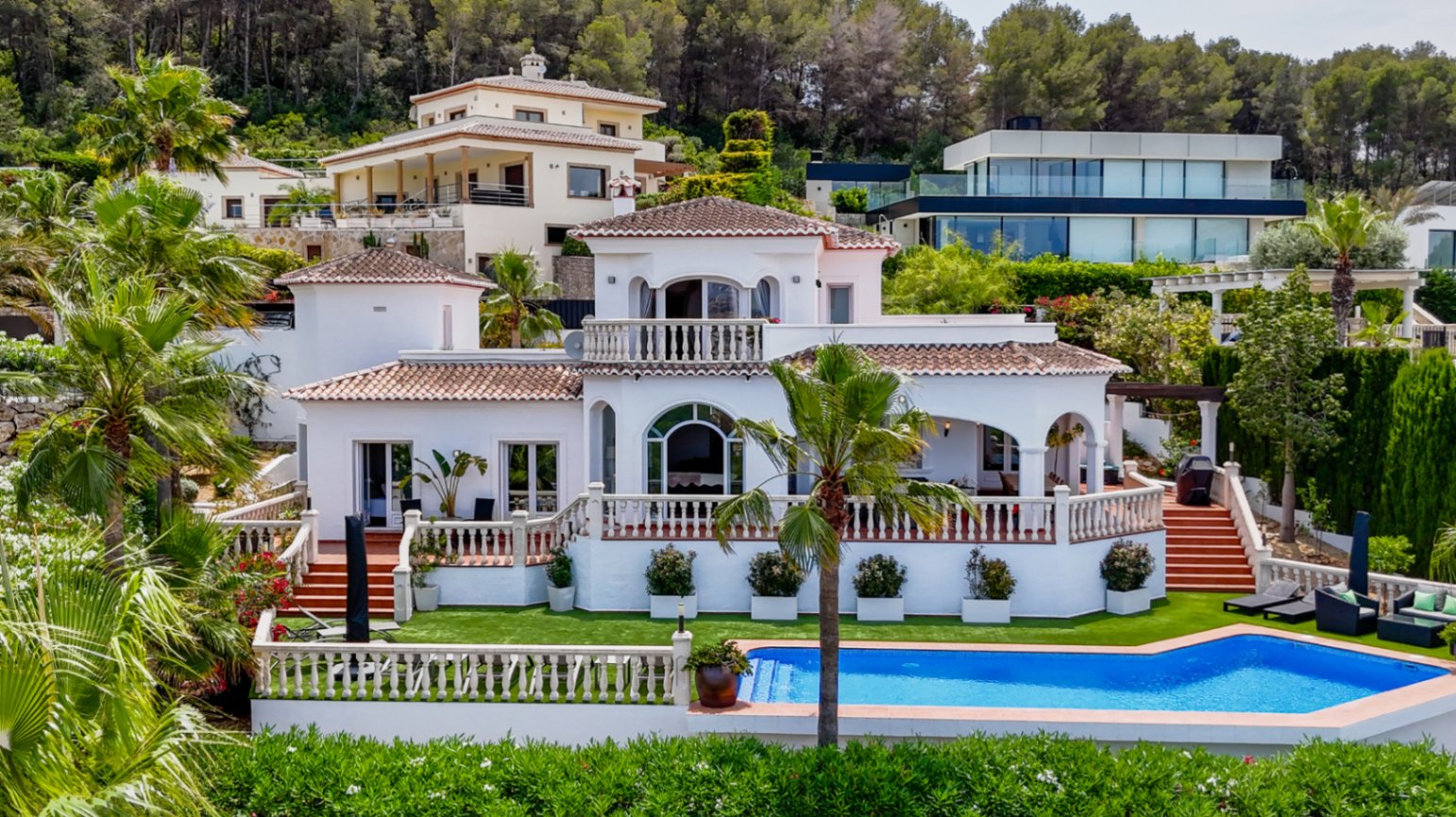 Mediterranean villa featuring a white facade, terracotta roofing, and tiered terraces with stone balustrades overlooking a private pool and palm garden.
