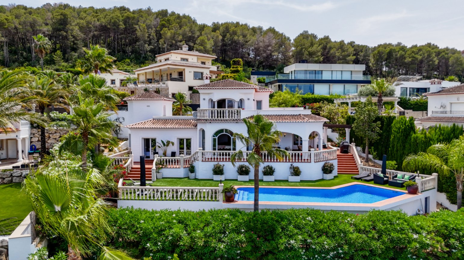 Mediterranean villa with white facade, terracotta roof, multi-level terraces, and a private pool surrounded by palm trees and pine-covered hills.