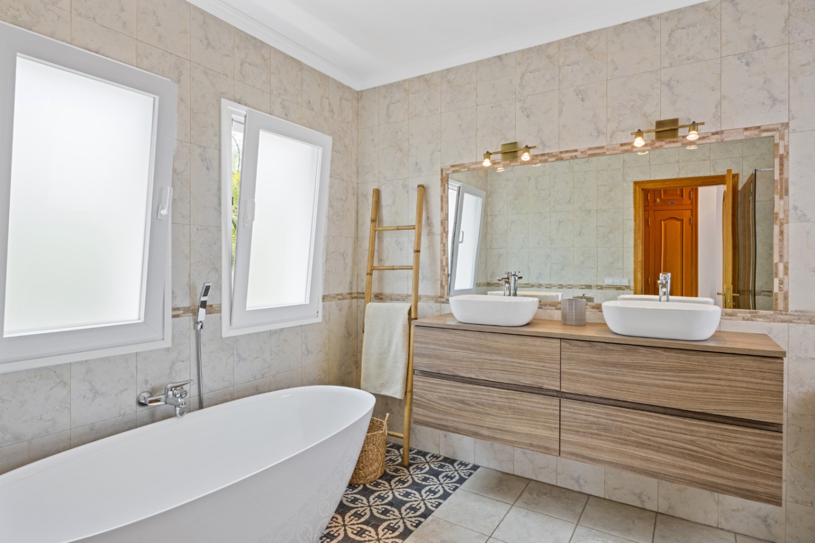 Modern bathroom featuring a freestanding bathtub, double wooden vanity with vessel sinks, and patterned floor tiles. Large mirror with brass fixtures and frosted windows.