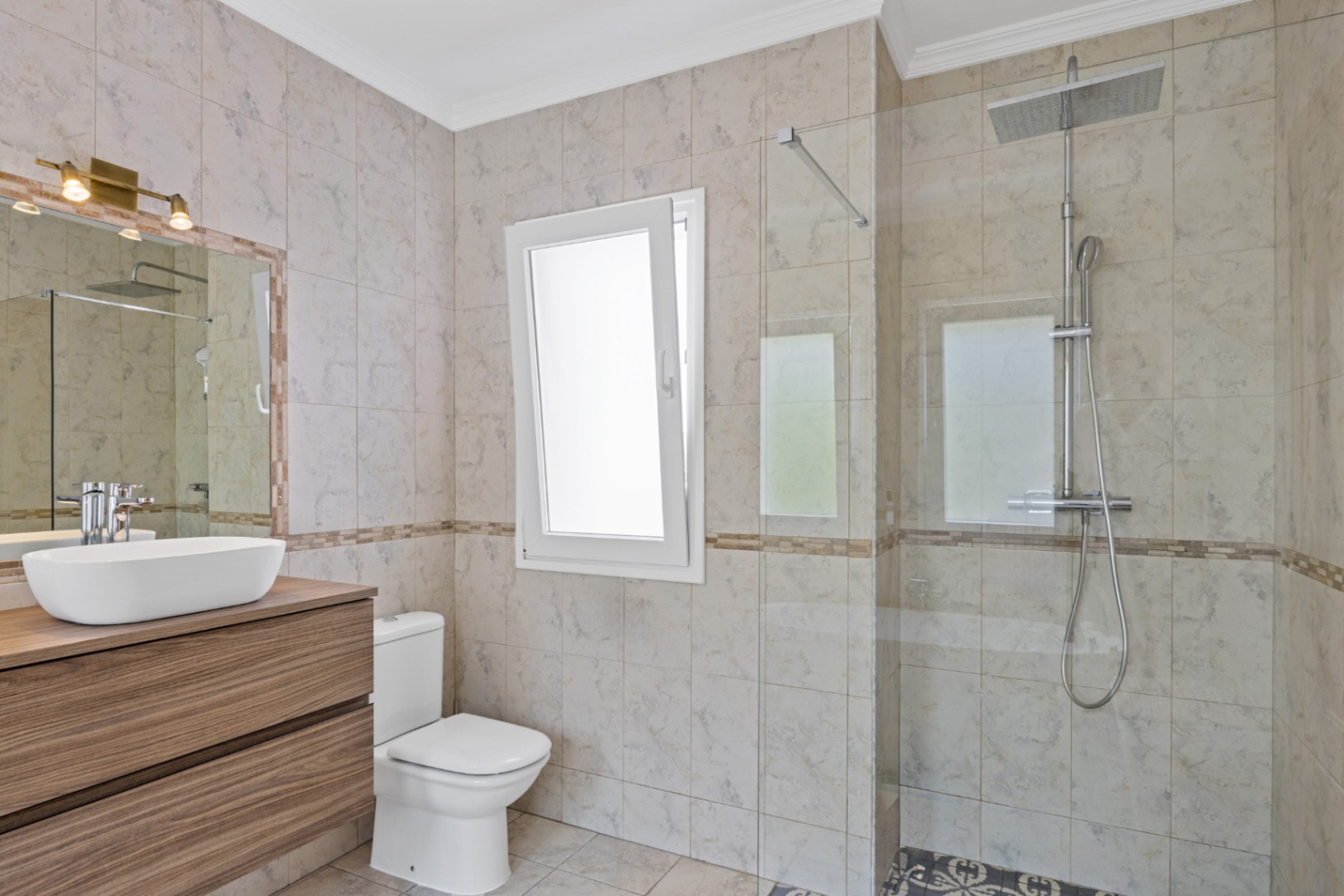 Modern bathroom featuring a walk-in shower with glass partition, a floating wooden vanity with vessel sink, and beige marble-effect tiling with natural window light.