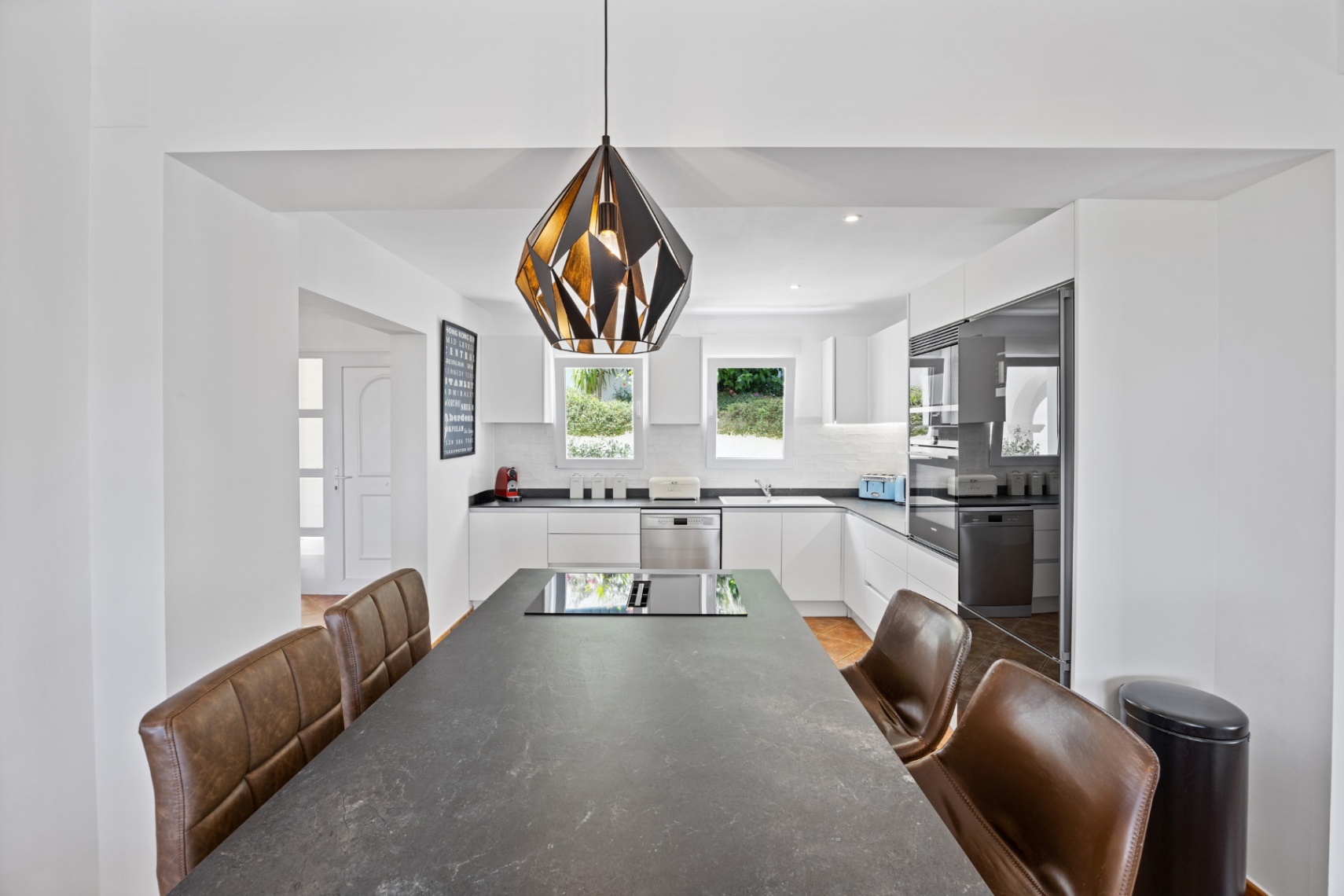 Modern kitchen featuring a large dark stone island with integrated induction hob, brown leather seating, and white cabinetry with built-in appliances.