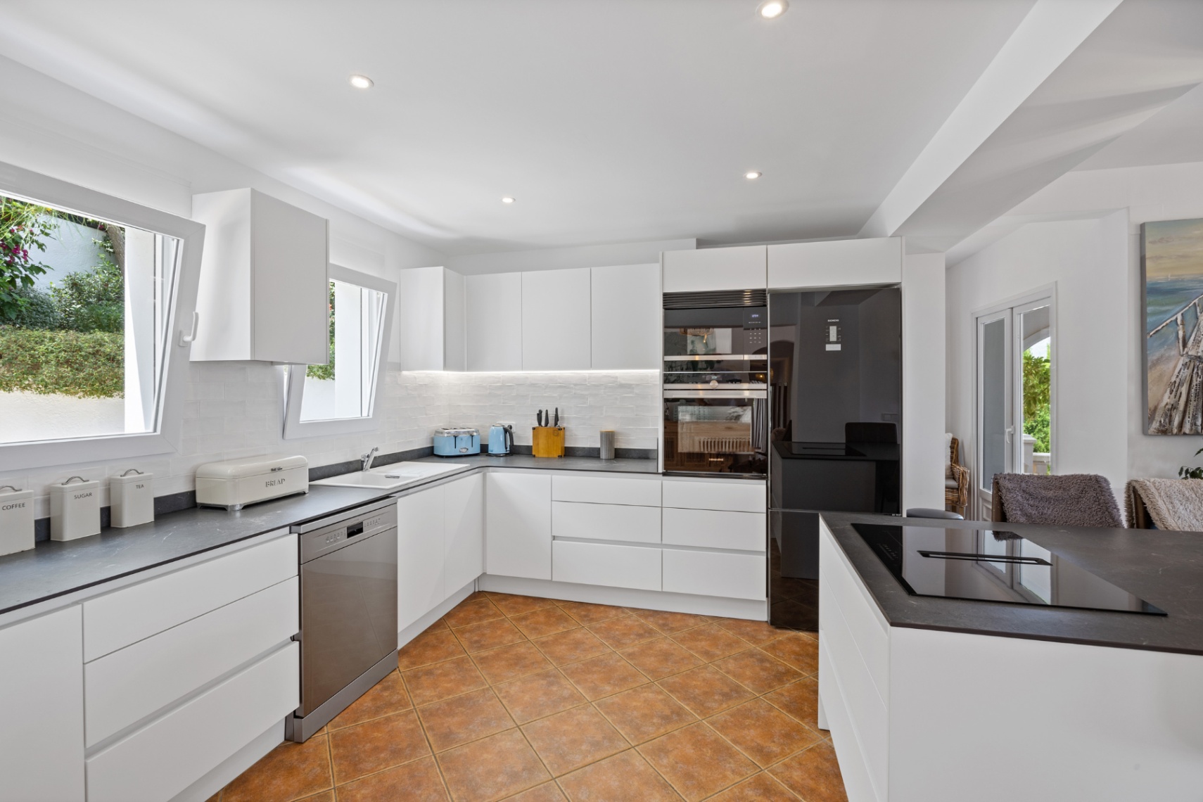 Modern kitchen featuring white handleless cabinetry, dark stone-effect countertops, integrated appliances, and terracotta-style ceramic floor tiles.