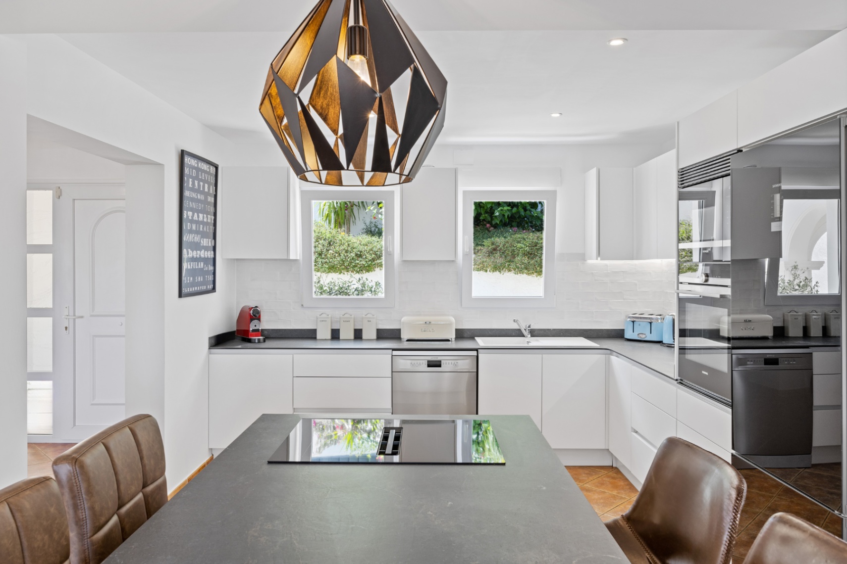 Modern kitchen with white handleless cabinetry, dark stone countertops, and a central island featuring an integrated cooktop and a striking geometric pendant light.