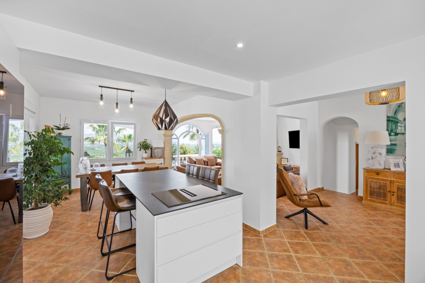 Open-plan kitchen featuring a white island with a dark countertop and integrated hob, terracotta tile flooring, and a dining area with views of palm trees.