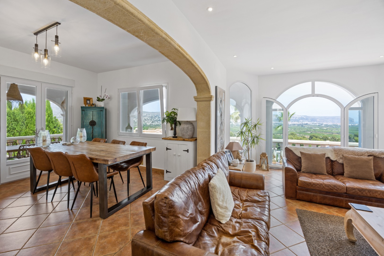 Open-plan living area featuring a stone arch, leather sofas, and a wooden dining table. Arched doors reveal panoramic valley views over terracotta tiles.