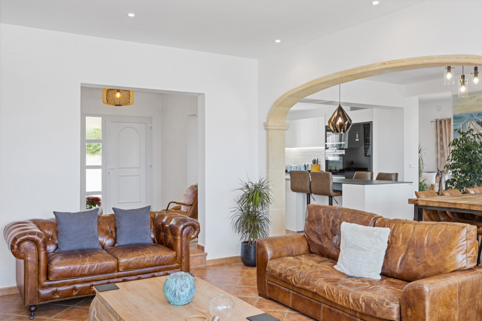 Open-plan living area featuring leather sofas, terracotta tile flooring, and a stone archway leading to a modern kitchen with a breakfast bar.