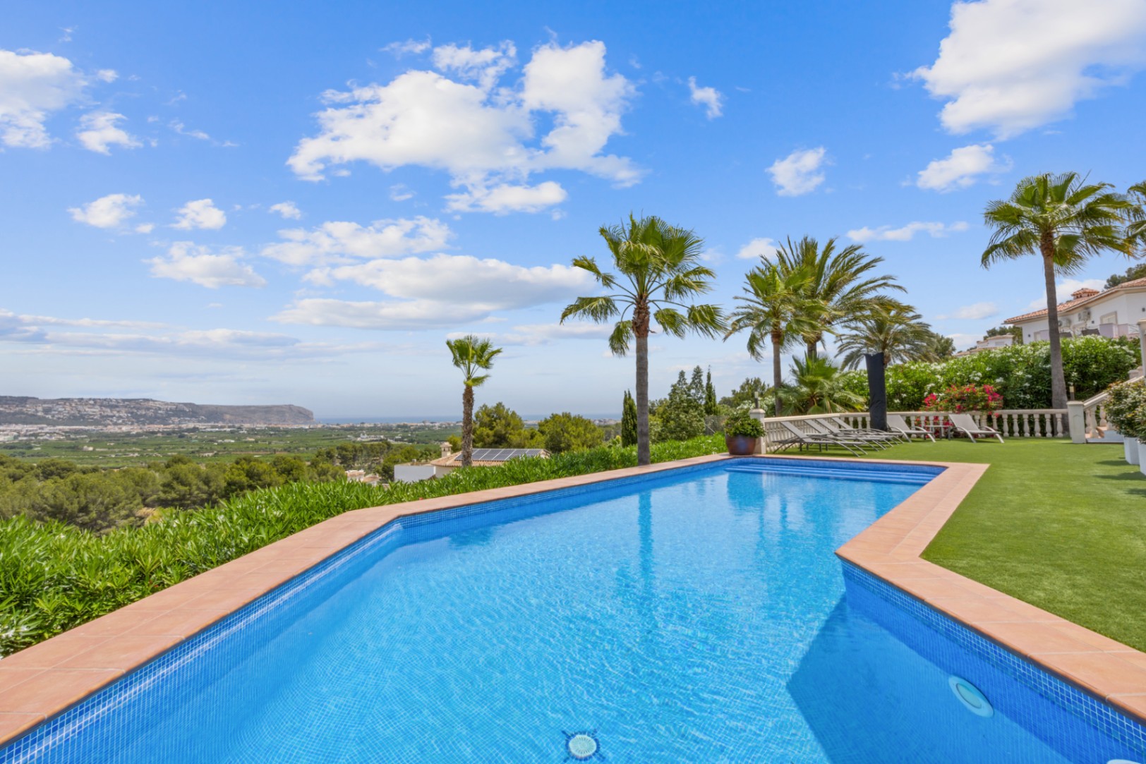 Outdoor swimming pool with blue mosaic tiling and terracotta coping, featuring panoramic views of the valley and sea, surrounded by palm trees and a lawn.