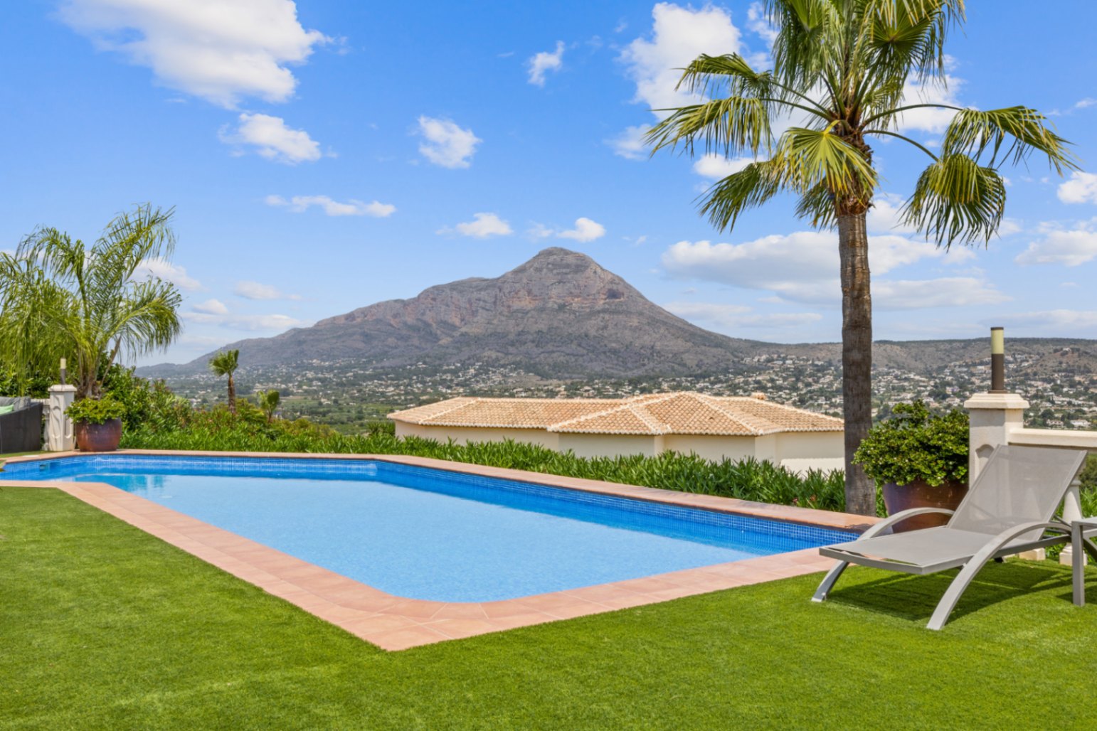 Outdoor swimming pool with blue mosaic tiling, surrounded by a synthetic lawn and terracotta border, offering panoramic views of a prominent mountain peak.