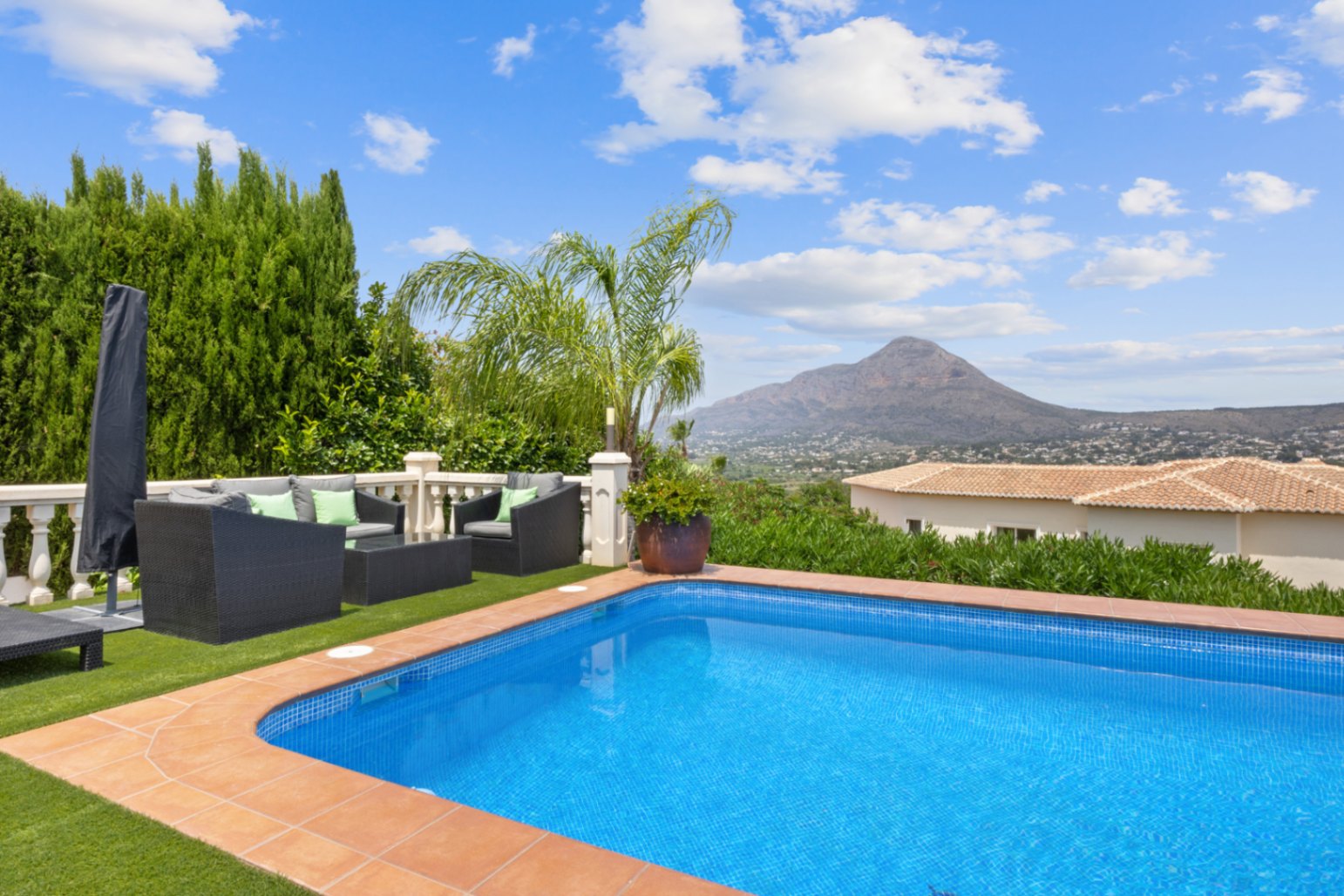 Outdoor swimming pool with terracotta tiling, a synthetic grass lounge area with wicker furniture, and panoramic views of the surrounding mountains.