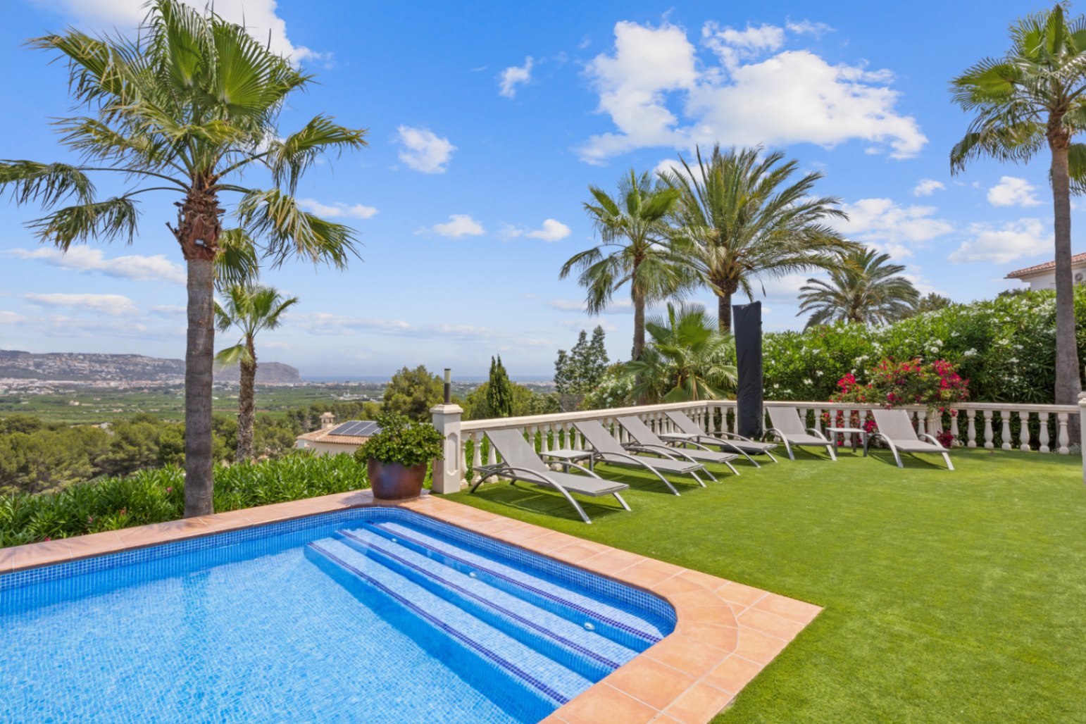 Poolside terrace featuring grey sun loungers on synthetic turf, a white stone balustrade, and panoramic views of the valley and Mediterranean coastline.