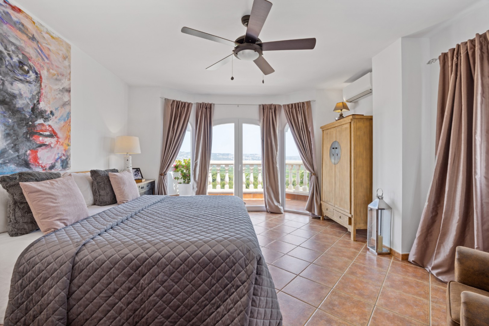 Spacious bedroom featuring terracotta flooring, a ceiling fan, and large arched doors opening onto a private balcony with scenic landscape views.