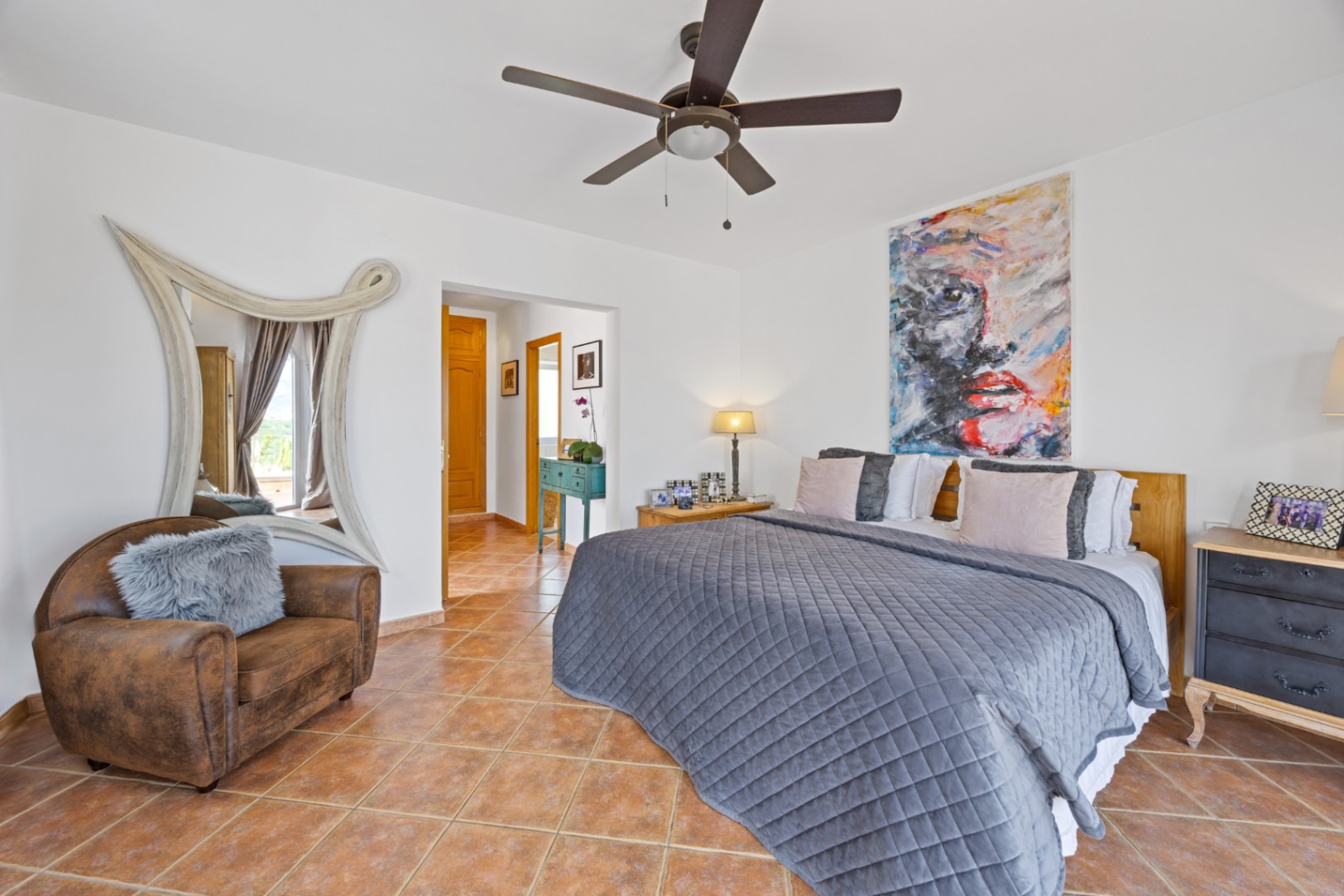 Spacious bedroom featuring terracotta tile floors, a large abstract portrait, and a decorative white-framed mirror. Includes a ceiling fan and a brown leather armchair.