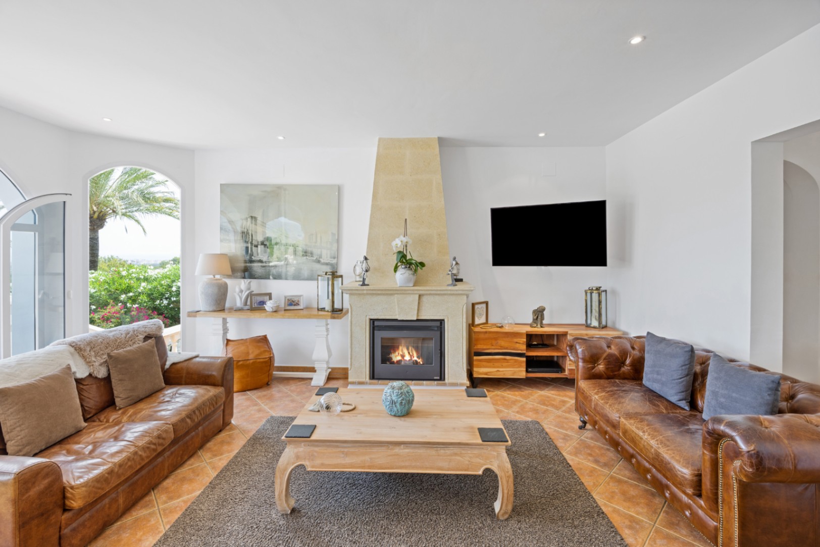 Spacious living area featuring a stone fireplace, brown leather sofas, and terracotta flooring. Large arched windows offer direct views of the Mediterranean garden.