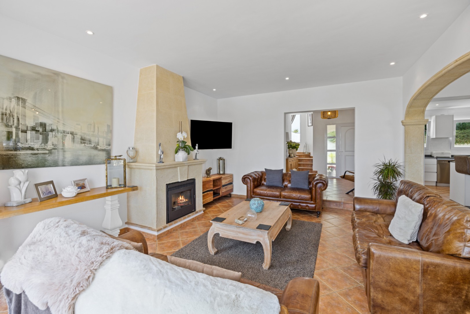 Spacious living room featuring brown leather sofas, a traditional stone fireplace, and terracotta tile flooring. An elegant stone archway leads to the kitchen.