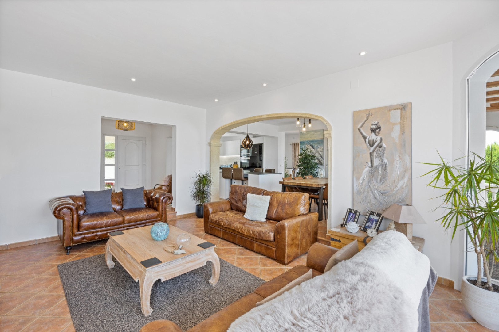 Spacious living room featuring brown leather sofas, terracotta tile flooring, and a stone-trimmed archway leading to the dining area and kitchen.