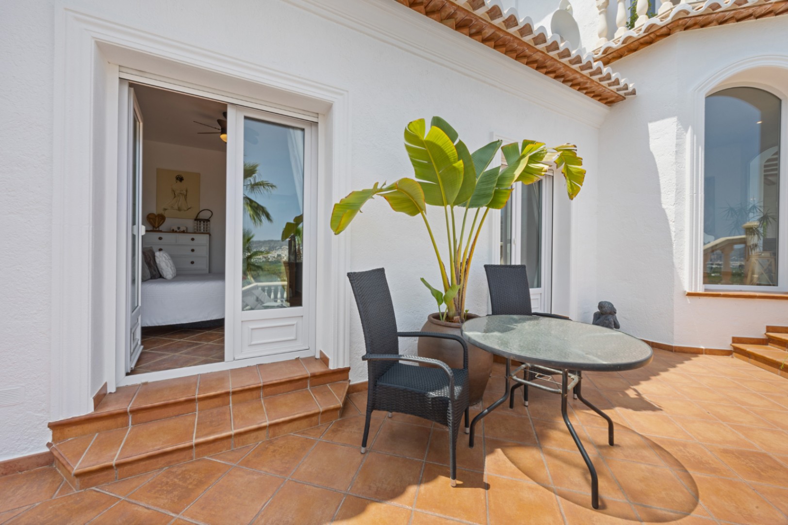 Terrace with terracotta tiling, outdoor seating, and a large potted plant, providing direct access to a bright bedroom through sliding glass doors.