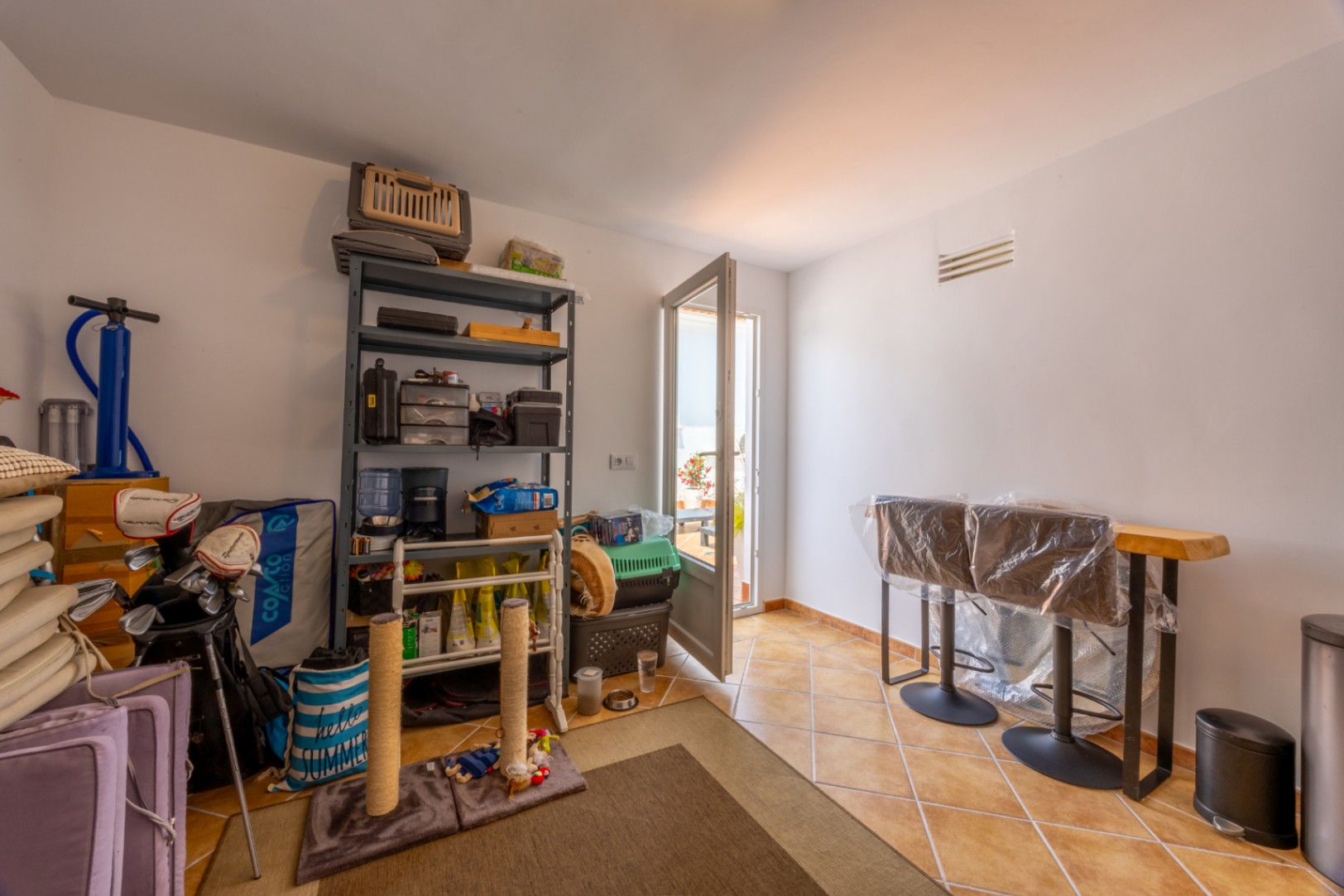 Utility room with terracotta tile flooring, featuring metal storage shelving, a wooden bar table with stools, and access to an outdoor terrace.