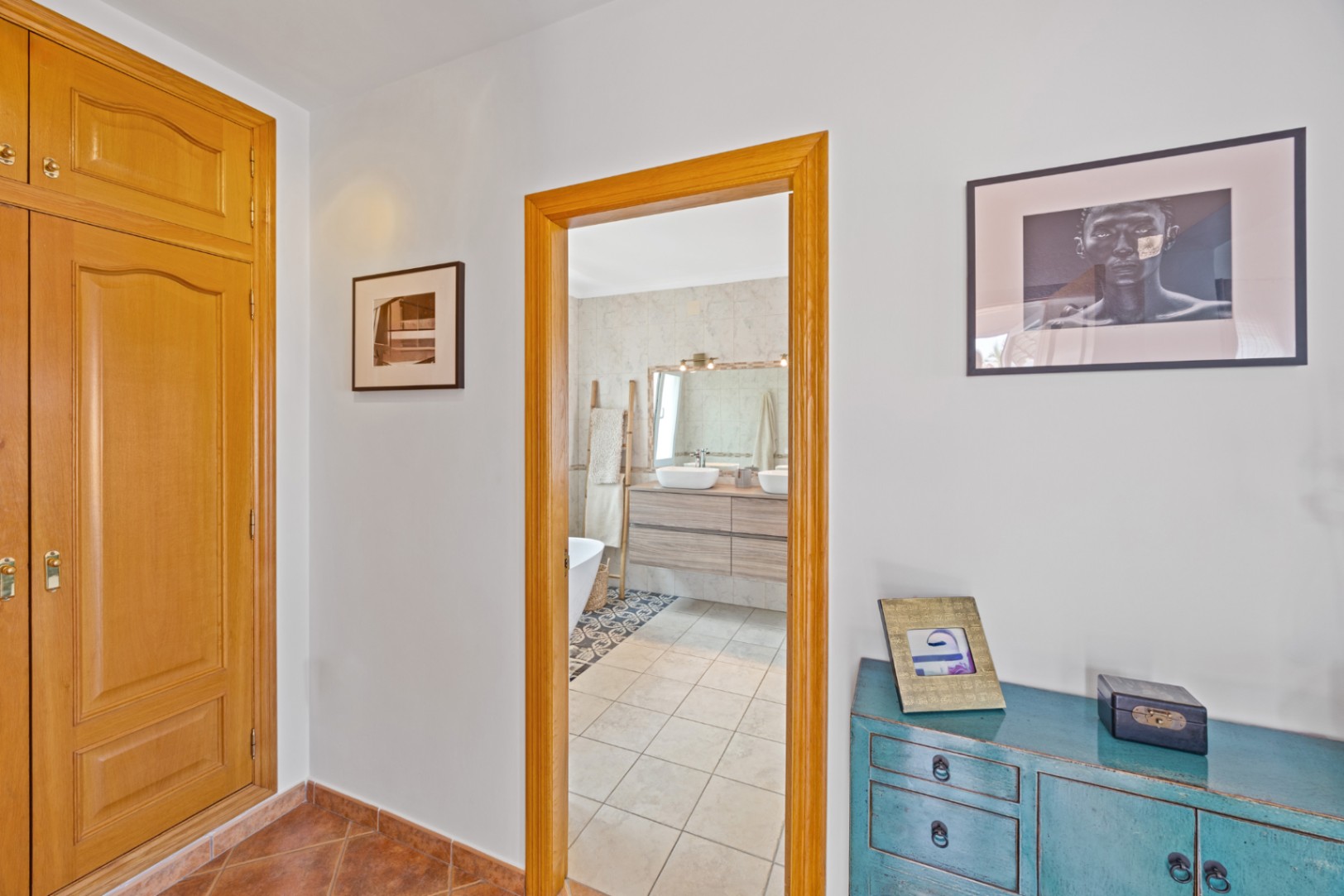 View from a hallway with built-in wooden wardrobes and a blue sideboard, looking into a bathroom featuring double vessel sinks and tiled finishes.