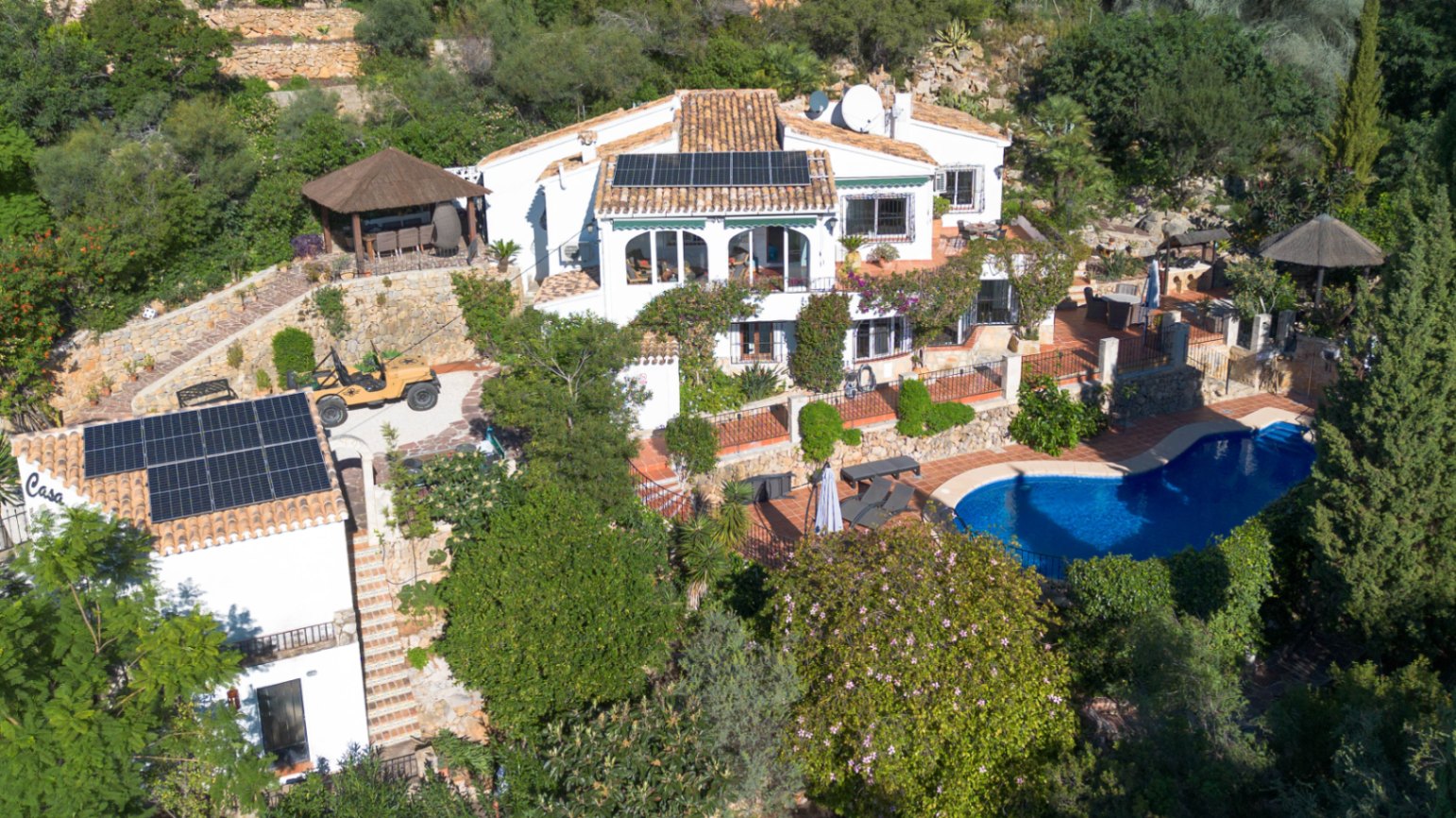 Aerial view of a Mediterranean villa featuring solar-paneled terracotta roofs, a kidney-shaped pool, and terraced gardens with stone walls and lush greenery.